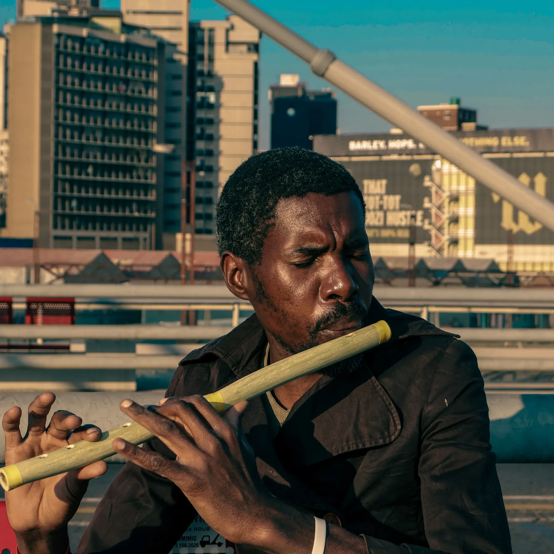 A man playing a hand-carved flute on a bridge, the Johannesburg skyline behind him.
