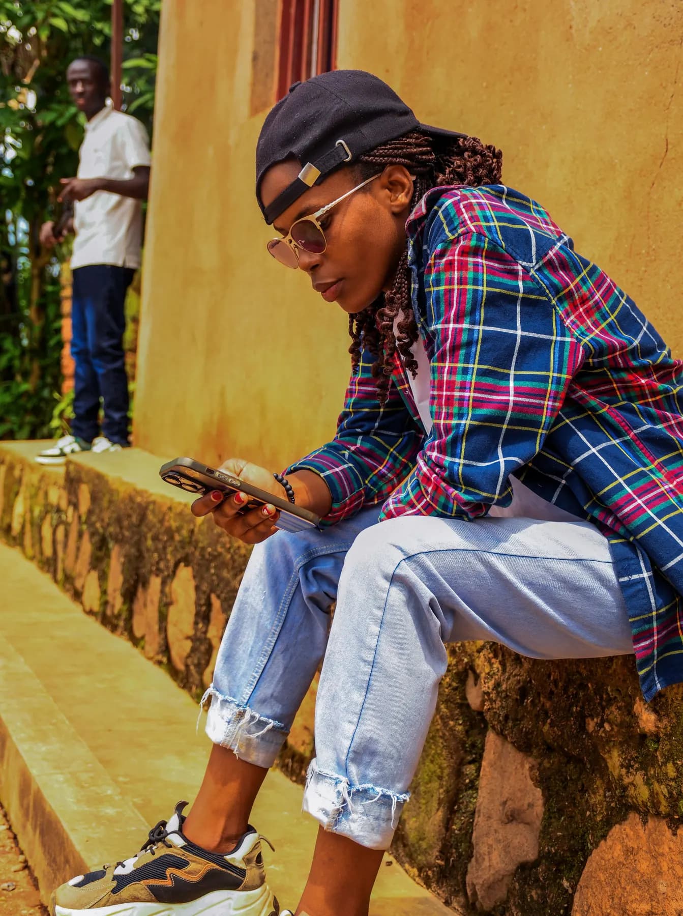 A young woman sitting on a low wall in the sun, looking at her phone.